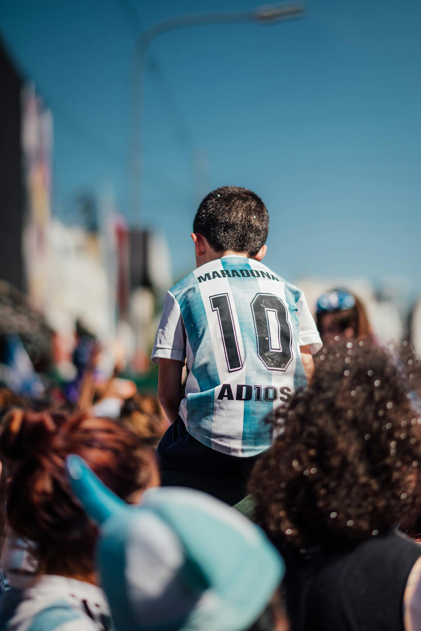 A child wearing a Maradona jersey sits on shoulders in a crowded Buenos Aires street, capturing the vibrant city atmosphere.